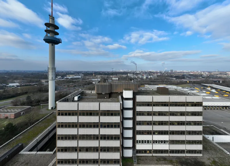 Drone shot at roof level: the high-rise before the retrofit, a telecom tower on the left, the city behind